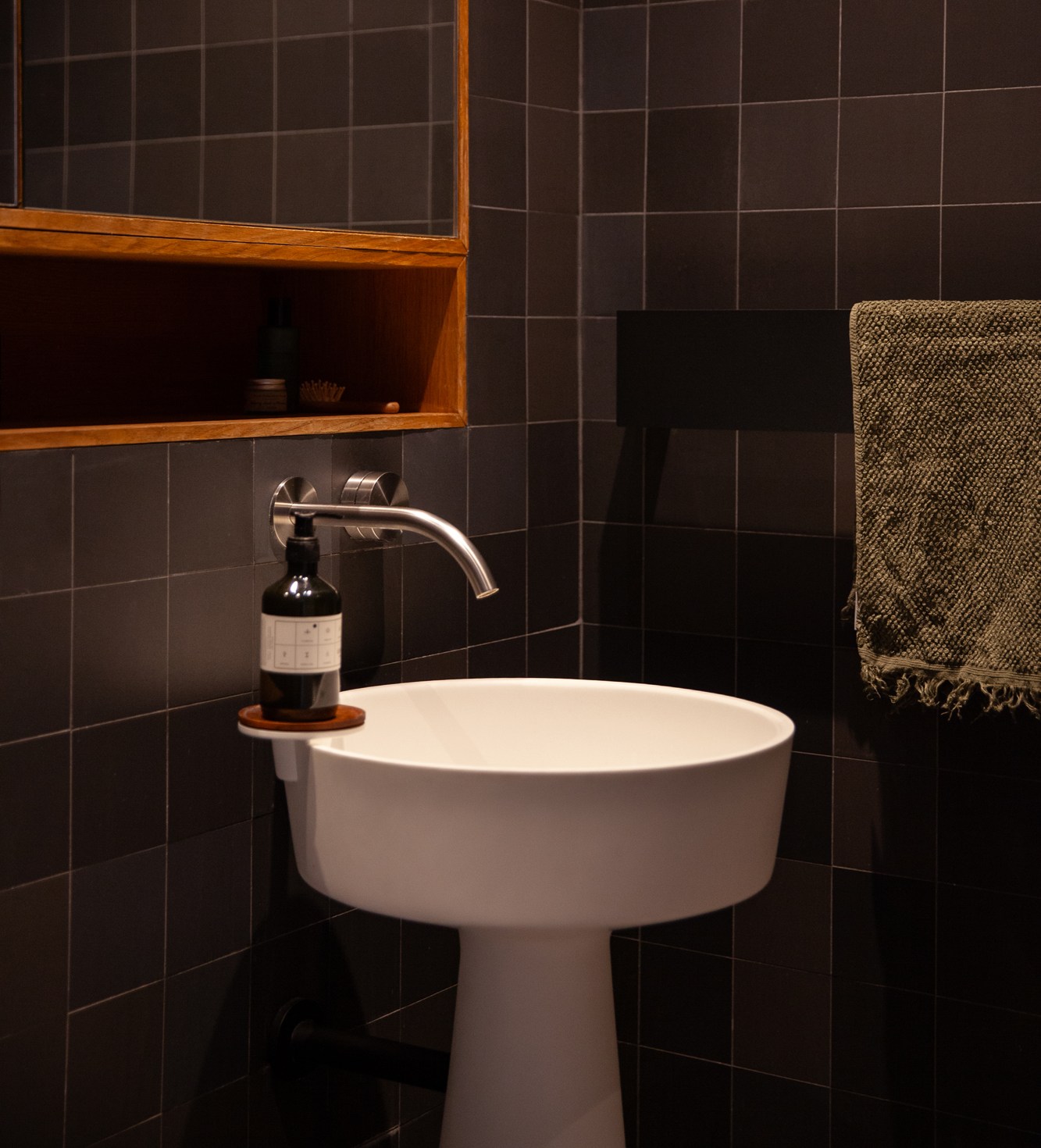 Bathroom interior with dark tiled walls, a white pedestal sink, a wooden mirrored cabinet, and a dark green towel.