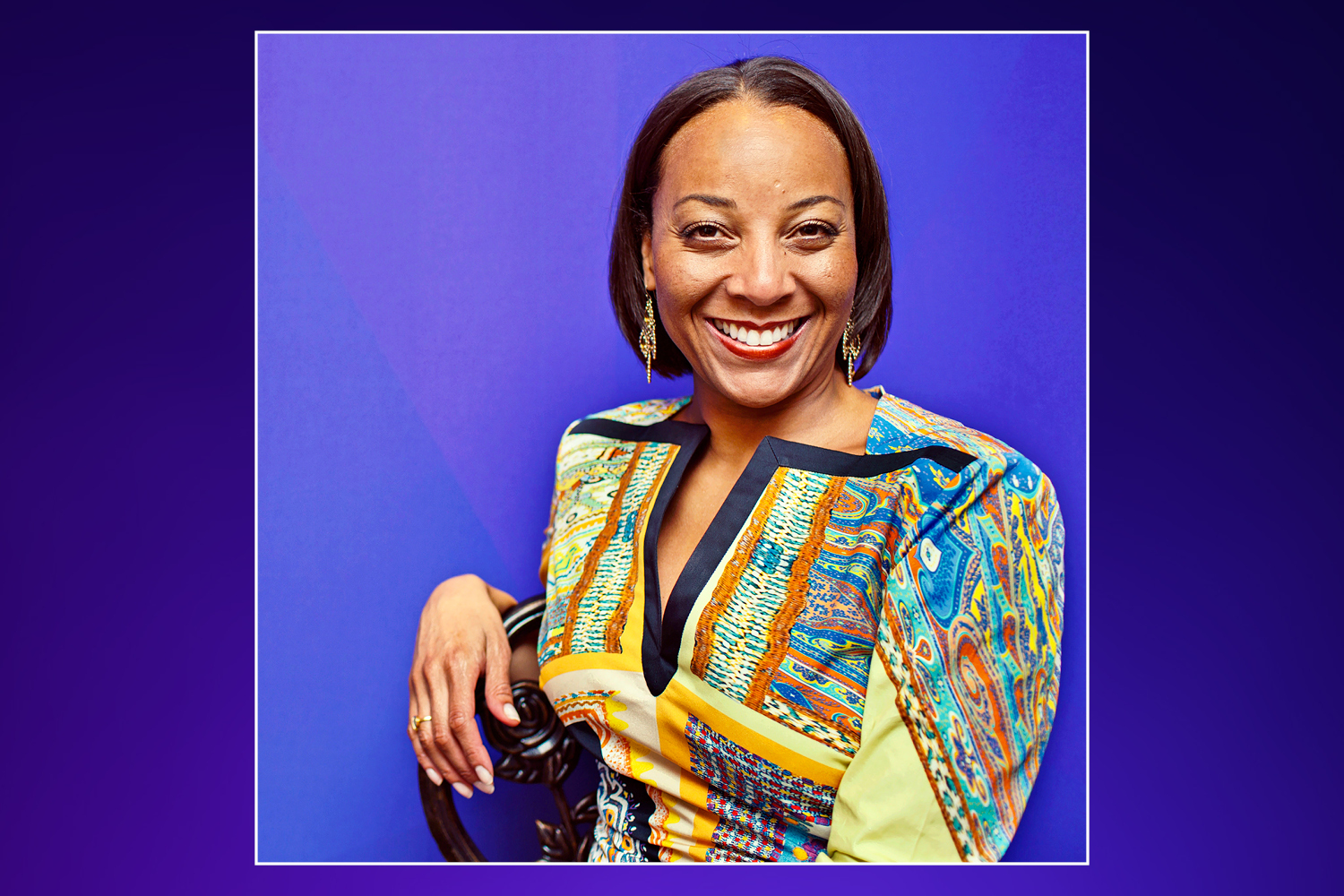 A smiling woman in a colorful patterned shirt sits in front of a blue background.