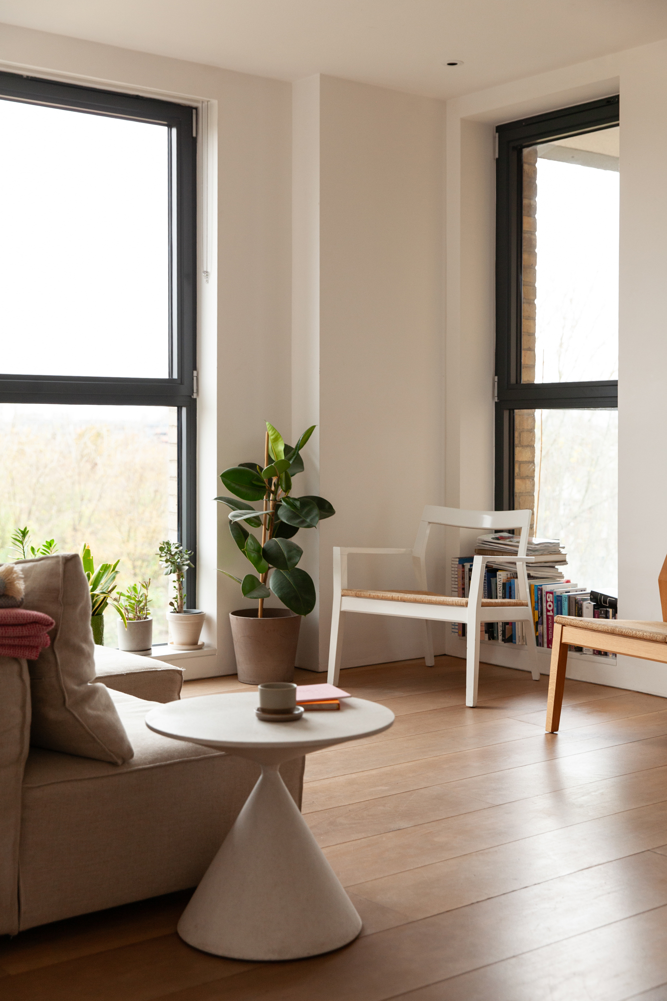 A minimalist living room with large windows, potted plants, a beige sofa, and a conical side table.