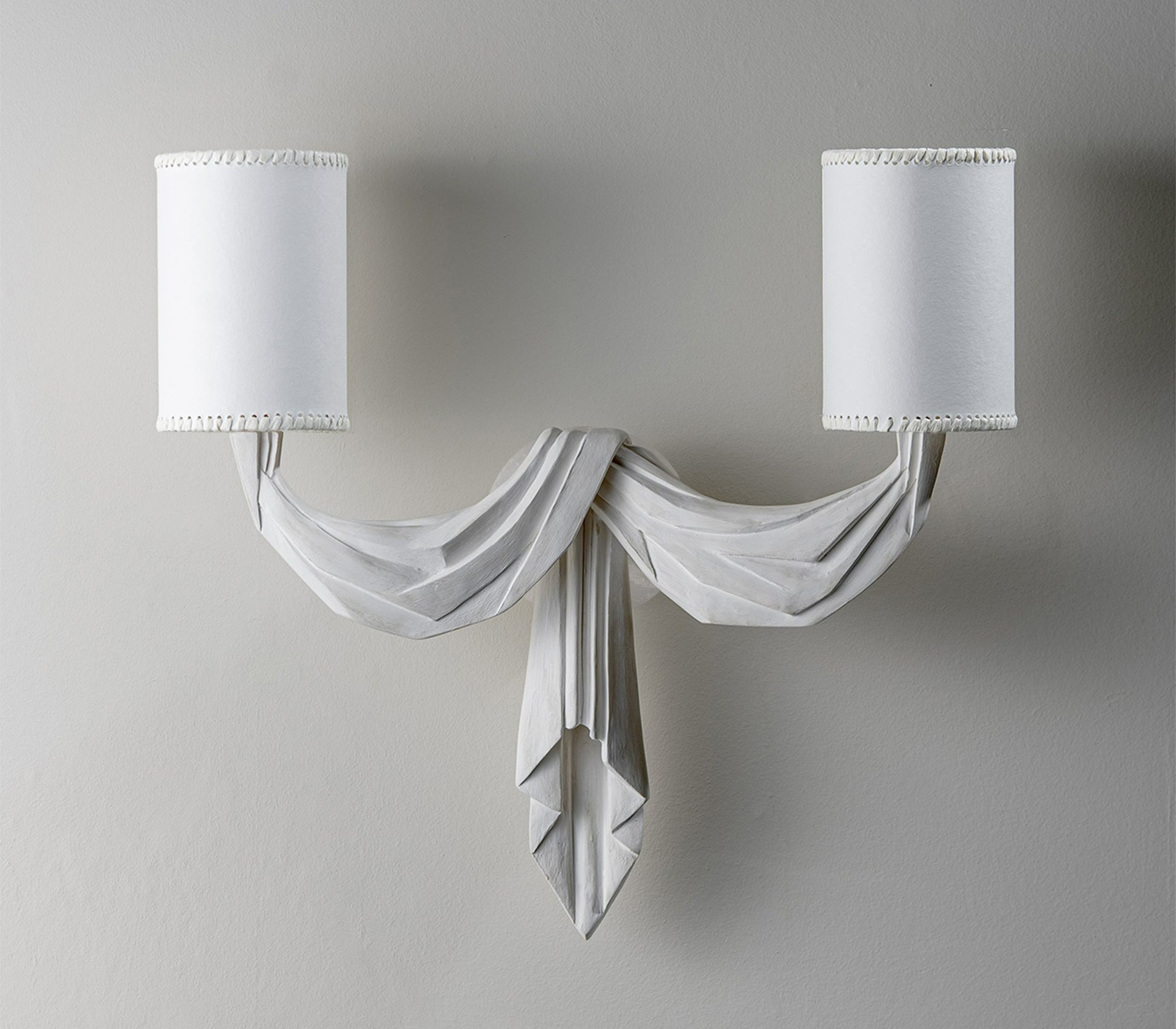 A white two-lamp wall sconce with a decorative draped centerpiece, resembling folded fabric.