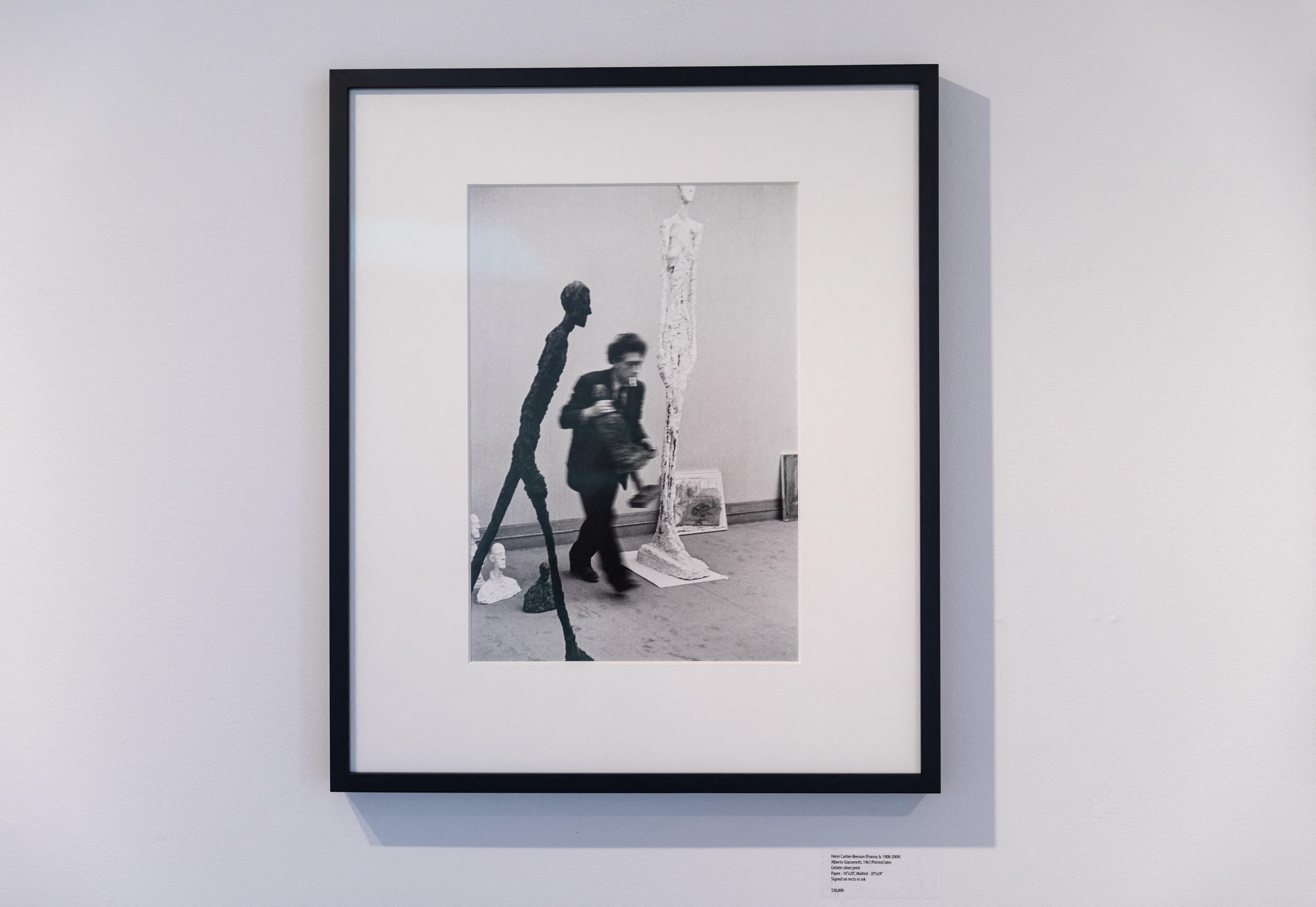 Black and white photo by Henri Cartier-Bresson showing Alberto Giacometti walking past his sculptures.