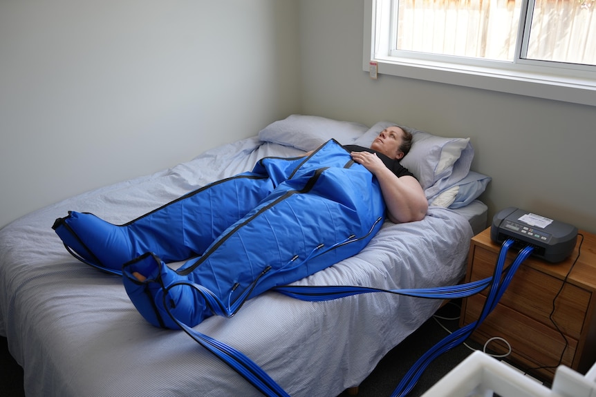 A photo showing a woman laying on the bed wearing a blue medical suit which is attached to a machine on a table.