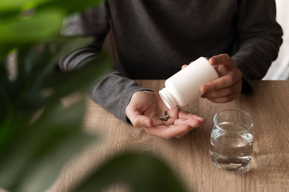 Adult man pouring supplement pills from a white bottle into his hand with a glass of water