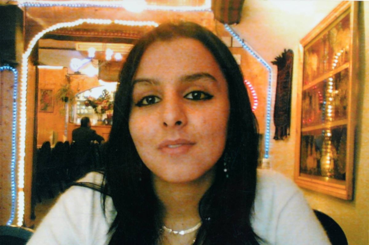 An individual with long dark hair, wearing a white top and a necklace, is seated in a room with a blurred background that includes an arched doorway, ambient lighting, and a wall adorned with framed artwork.