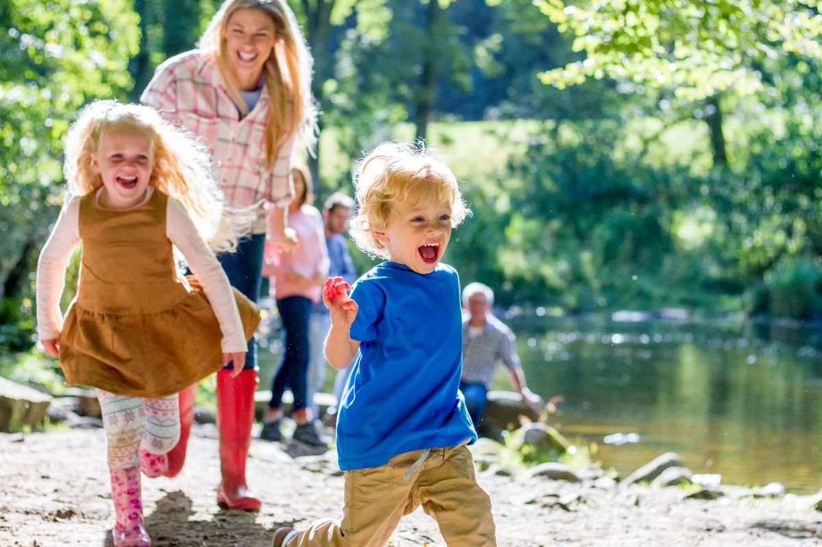 Children joyfully run along a river's edge, with a woman and other individuals in the background, amidst a lush, green setting.