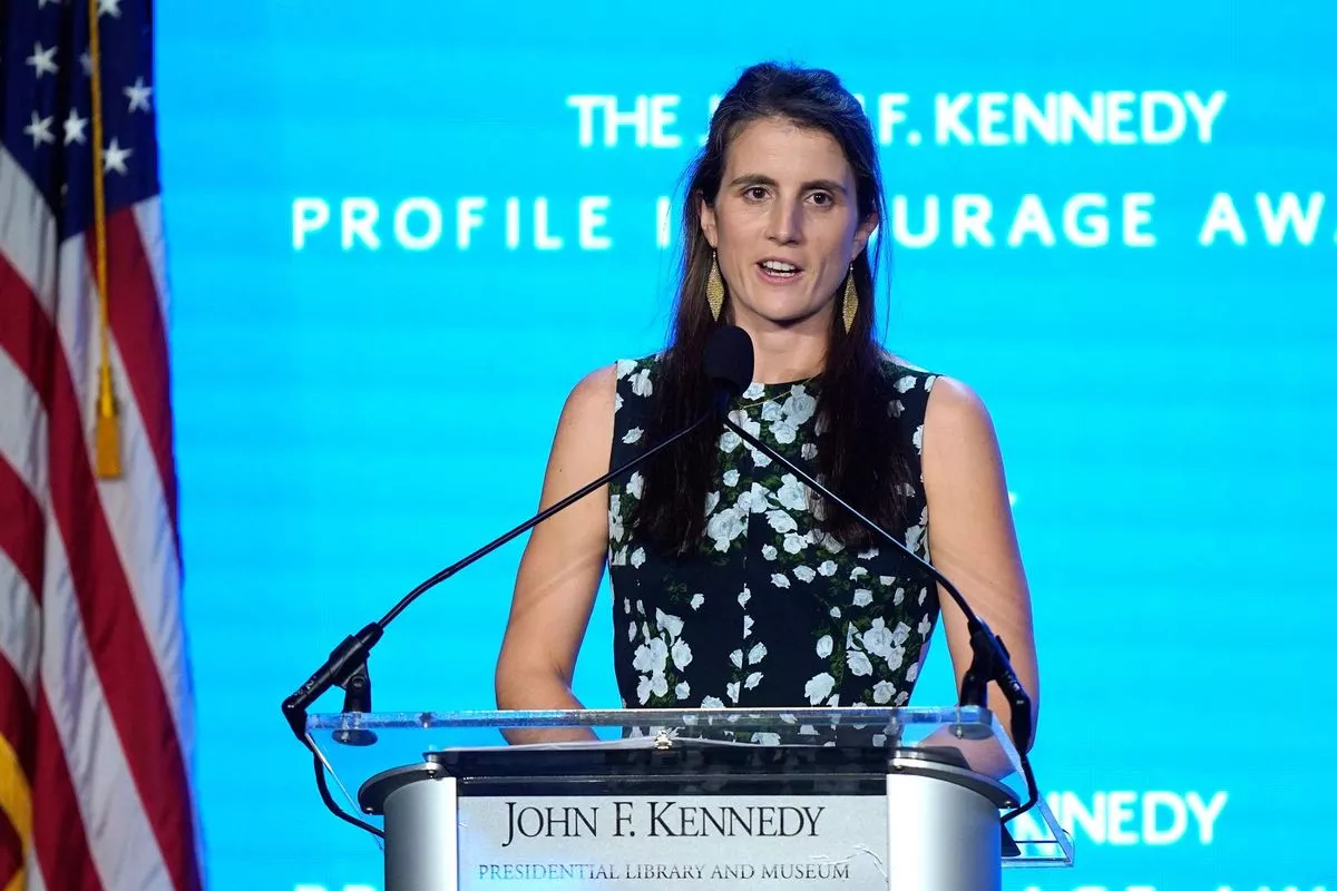 An individual with long hair, wearing a floral patterned top, is standing in front of a microphone and speaking at an event. Behind them, a blue backdrop displays the words "The F. Kennedy" and "ILE I URAC."