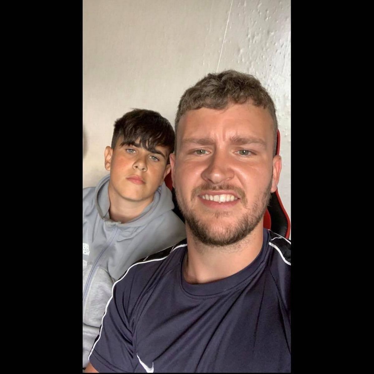 Two individuals are standing close together, one with his eyes closed and the other with a broad smile, against a plain background.