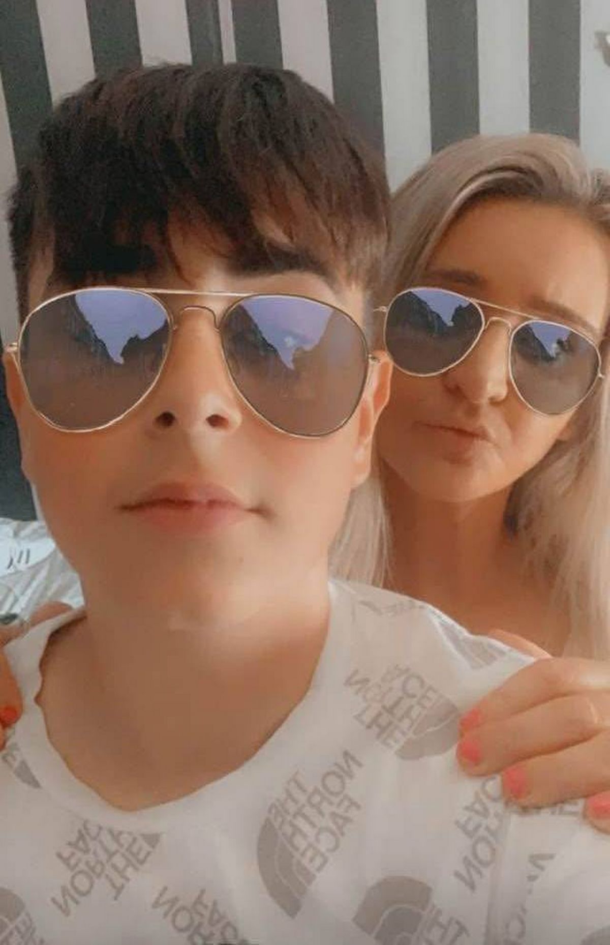 Two individuals, one male and one female, are both wearing aviator sunglasses with reflective lenses. The male subject is looking directly at the camera while the female subject is positioned slightly behind and to the side, also gazing towards the camera.