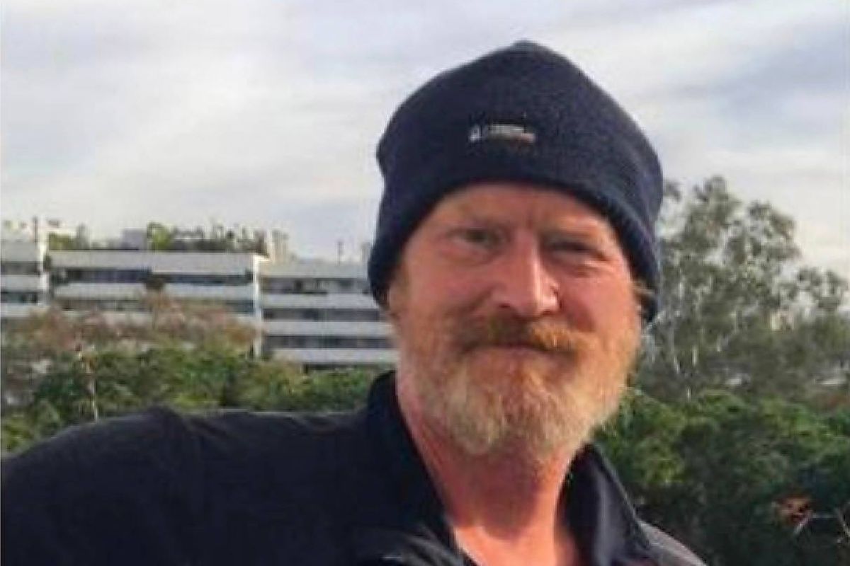 An individual with a beard and mustache, donning a navy blue beanie, is captured in a photograph against a backdrop that includes a building and greenery.