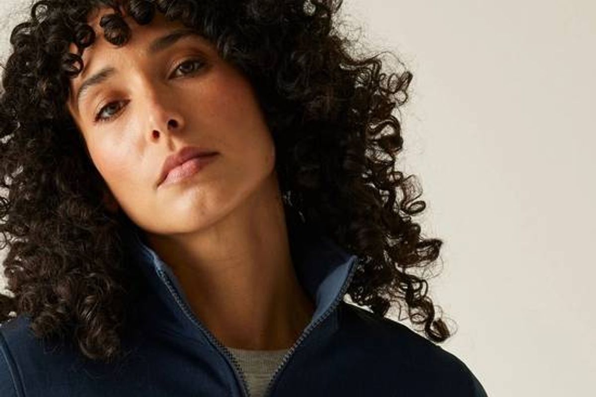 An individual with curly hair, wearing a navy blue fleece jacket, is standing against a plain background and appears to be posing for a photograph.