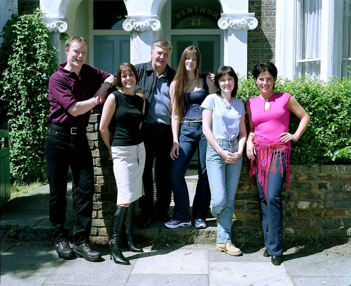 A group of individuals, dressed in a variety of contemporary attire, standing together in front of a building with a backdrop of greenery.
