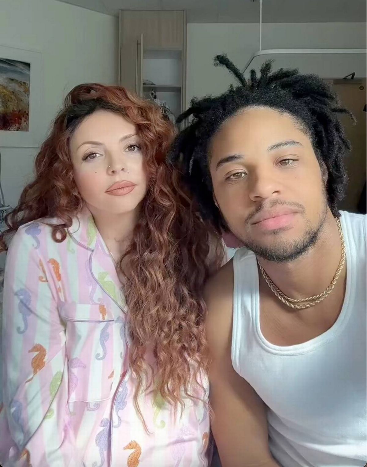 An image depicts a man and a woman standing close together, posing for a photograph. The woman, with long flowing hair, is wearing a pink top and appears to be smiling. The man, with a beard and dreadlocks, is wearing a white sleeveless top and has a contemplative expression. They both appear to be in a casual setting, with a wall and a plant visible in the background.