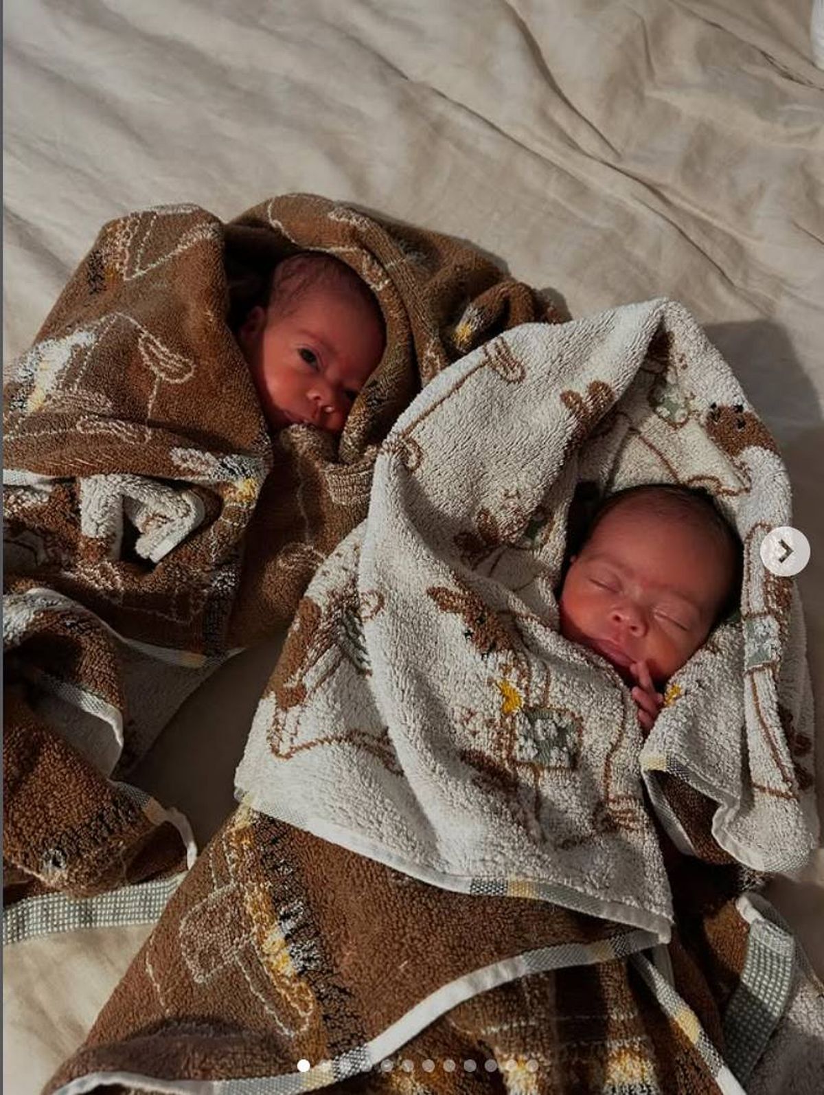 Two infants, wrapped in camouflage-patterned blankets, are peacefully sleeping side by side. One of the infants has its head slightly turned to the side, while the other rests with its head facing forward.