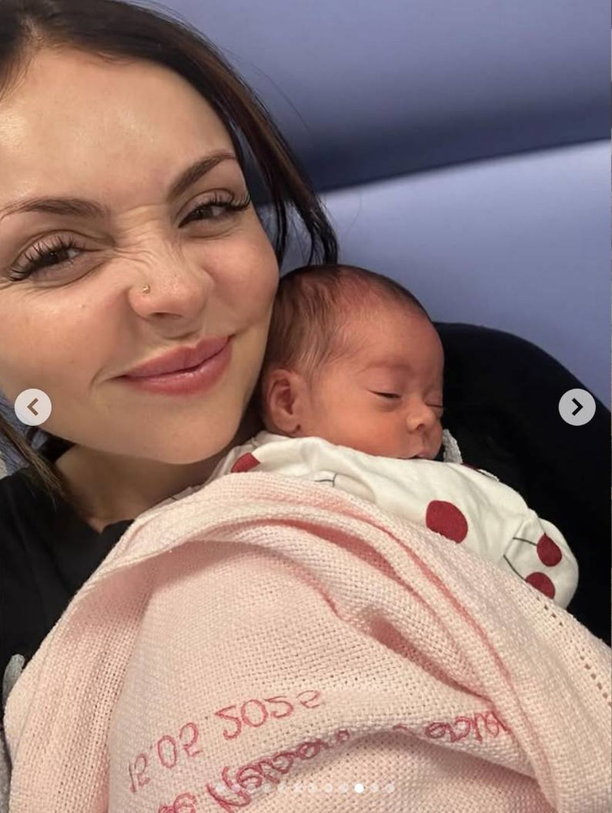 An individual, presumably a mother, is tenderly holding a newborn infant in her arms, both displaying serene expressions. The mother is dressed in a dark top, and the infant is wrapped in a white garment. The setting appears to be a cozy, intimate environment.