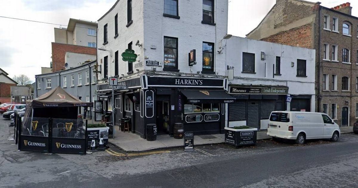 An urban street scene featuring a large white building with several windows and storefronts, partially shaded by awnings. A few vehicles are parked in front, including a white van and a dark-colored car. The sidewalk is adjacent to the building, and the area appears to be a commercial district with other buildings and structures in the background.