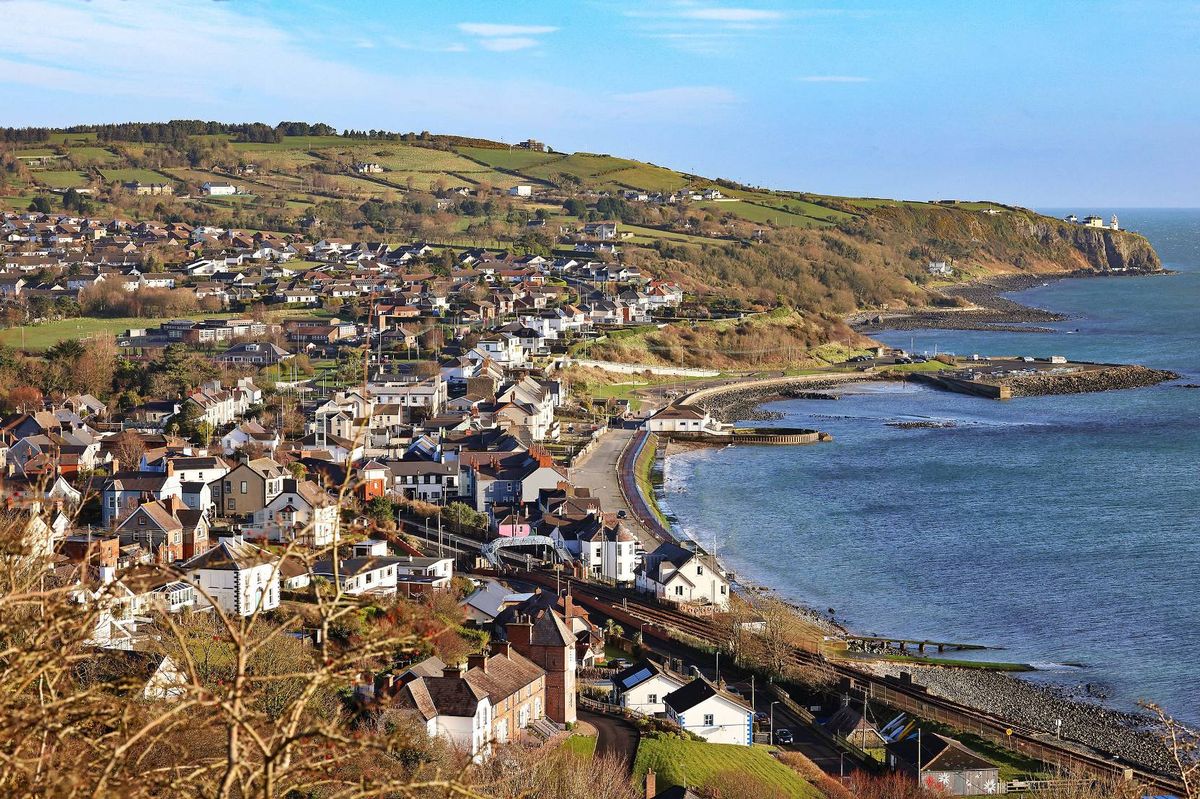 An aerial view of a coastal town situated at the base of a hill, with a clear blue sky overhead. The town features a collection of buildings interspersed with patches of greenery, and it is bordered by a calm body of water, possibly a sea or a large lake.