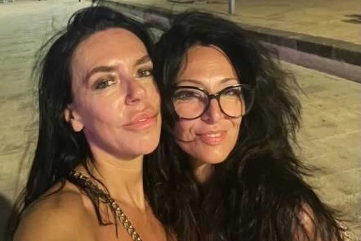 Two women with long black hair are smiling and posing closely together for a photograph in an outdoor setting.