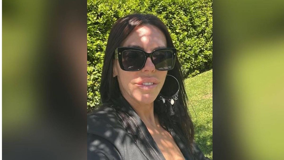 A woman with long hair, wearing sunglasses and a black jacket, is standing in an outdoor area with greenery in the background. She is smiling and appears to be posing for a photograph.