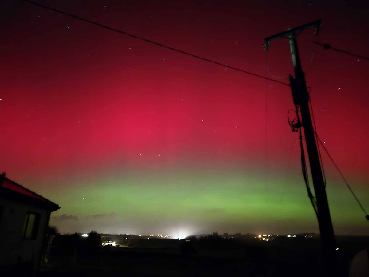 An image depicting the night sky with vivid auroral displays, characterized by a striking contrast between red and green hues against a dark backdrop, with a distant structure partially visible in the foreground.