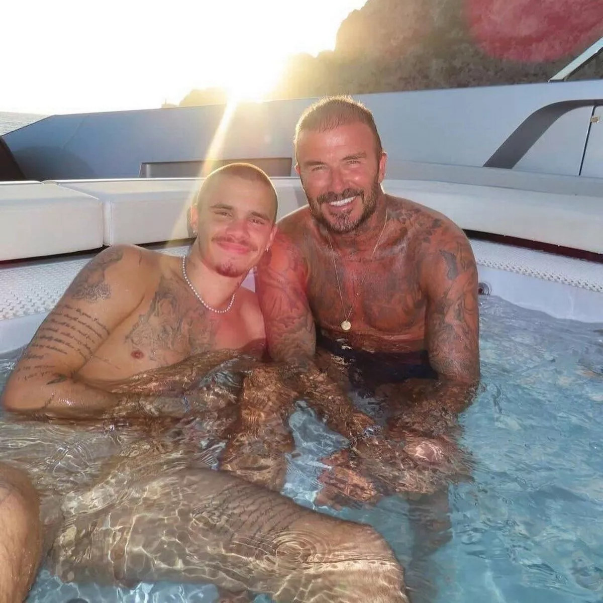 Two individuals, a man and a woman, are seated together in a jacuzzi, their bodies partially submerged in the water. They appear to be relaxed and are gazing in the same direction, possibly capturing a moment on camera. The setting suggests an outdoor environment, with a boat or deck visible in the background. The sun is setting in the distance, casting a warm glow over the scene.