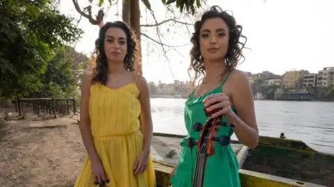 Celtic Connections The Ayoub sisters standing by a river, wearing dresses - one yellow, one green, and holding a violin and cello,