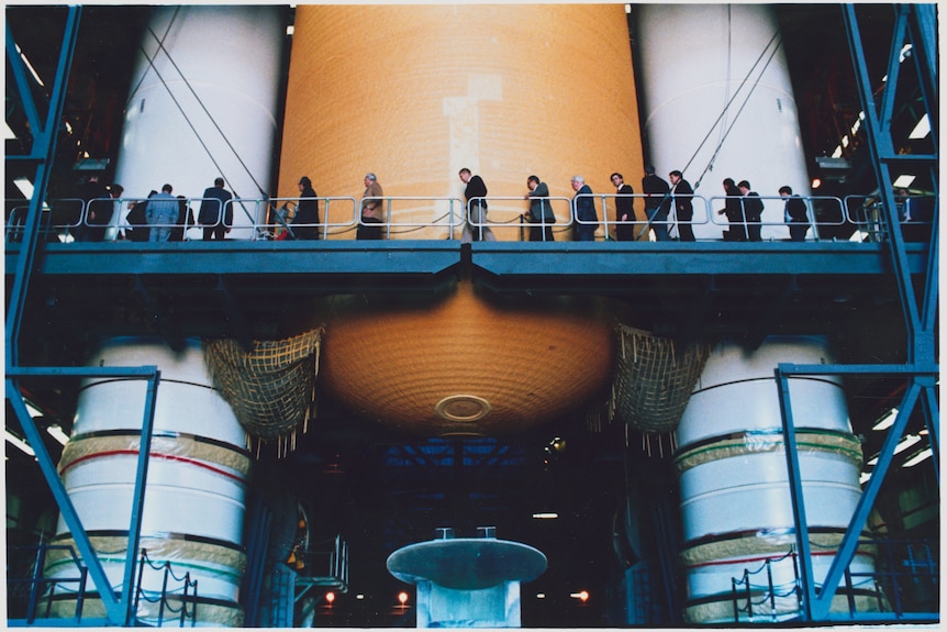 Wide shot of people dressed in suits crossing a high walkway, dwarfed by the a huge orange tank and two white rockets behind.