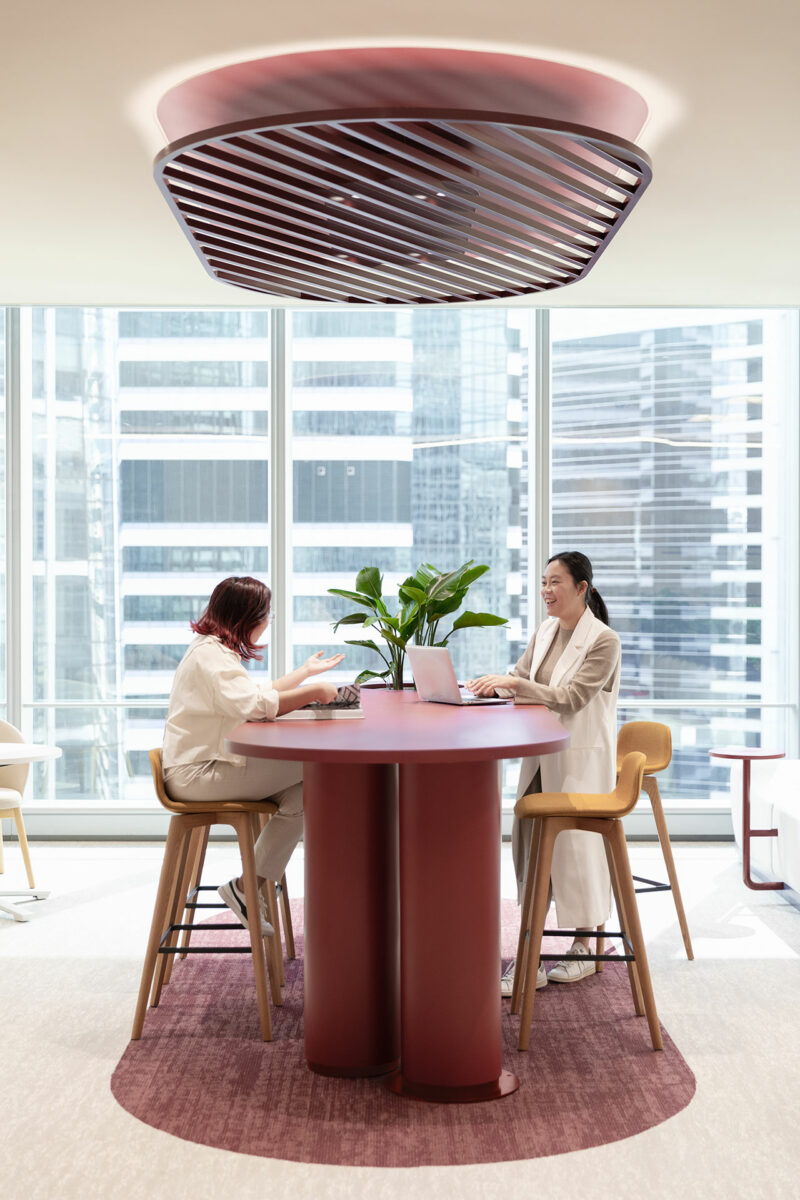 Two women work at a tall round red table in a modern office with large windows and city views, a plant centerpiece, and a geometric ceiling fixture above.