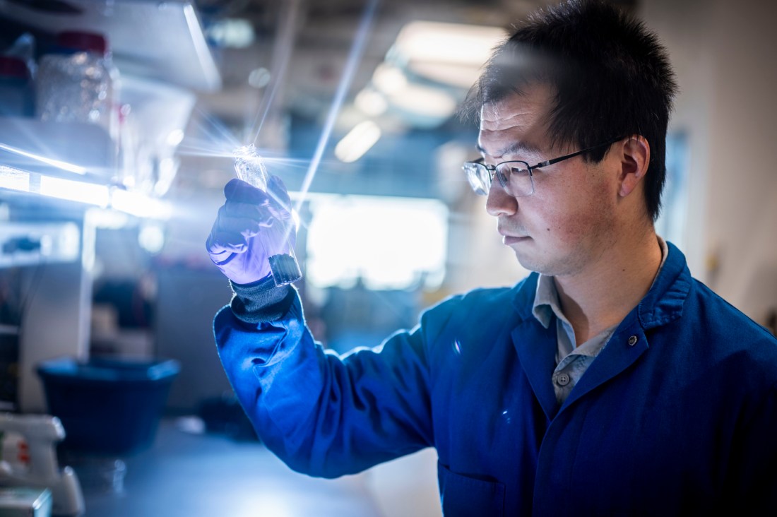 Zhenyu Tian, wearing a lab coat and gloves, holds up and inspects a test tube that contains crumb rubber.