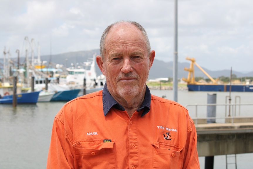A fisherman in high-vis clothing looking seriously at the camera with boats in the background.