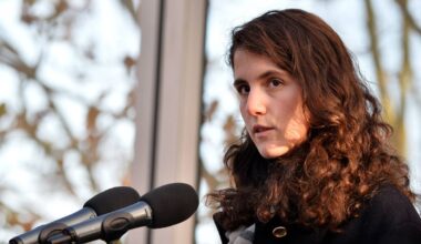 An individual with long hair, wearing a floral patterned top, is standing in front of a microphone and speaking at an event. Behind them, a blue backdrop displays the words "The F. Kennedy" and "ILE I URAC."
