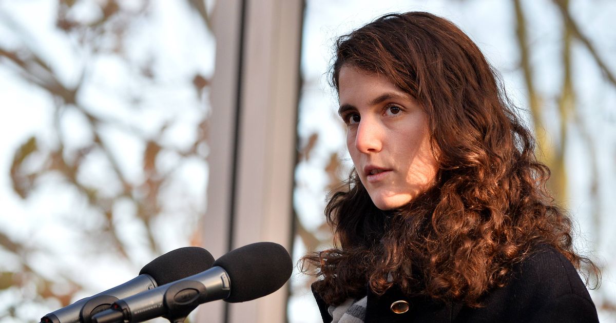 An individual with long hair, wearing a floral patterned top, is standing in front of a microphone and speaking at an event. Behind them, a blue backdrop displays the words "The F. Kennedy" and "ILE I URAC."