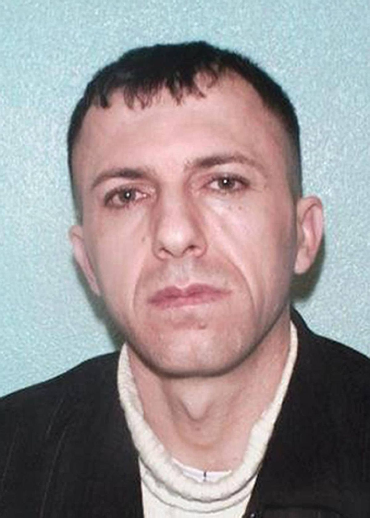 An individual with a serious expression, dressed formally, is shown in a close-up portrait against a light blue background.