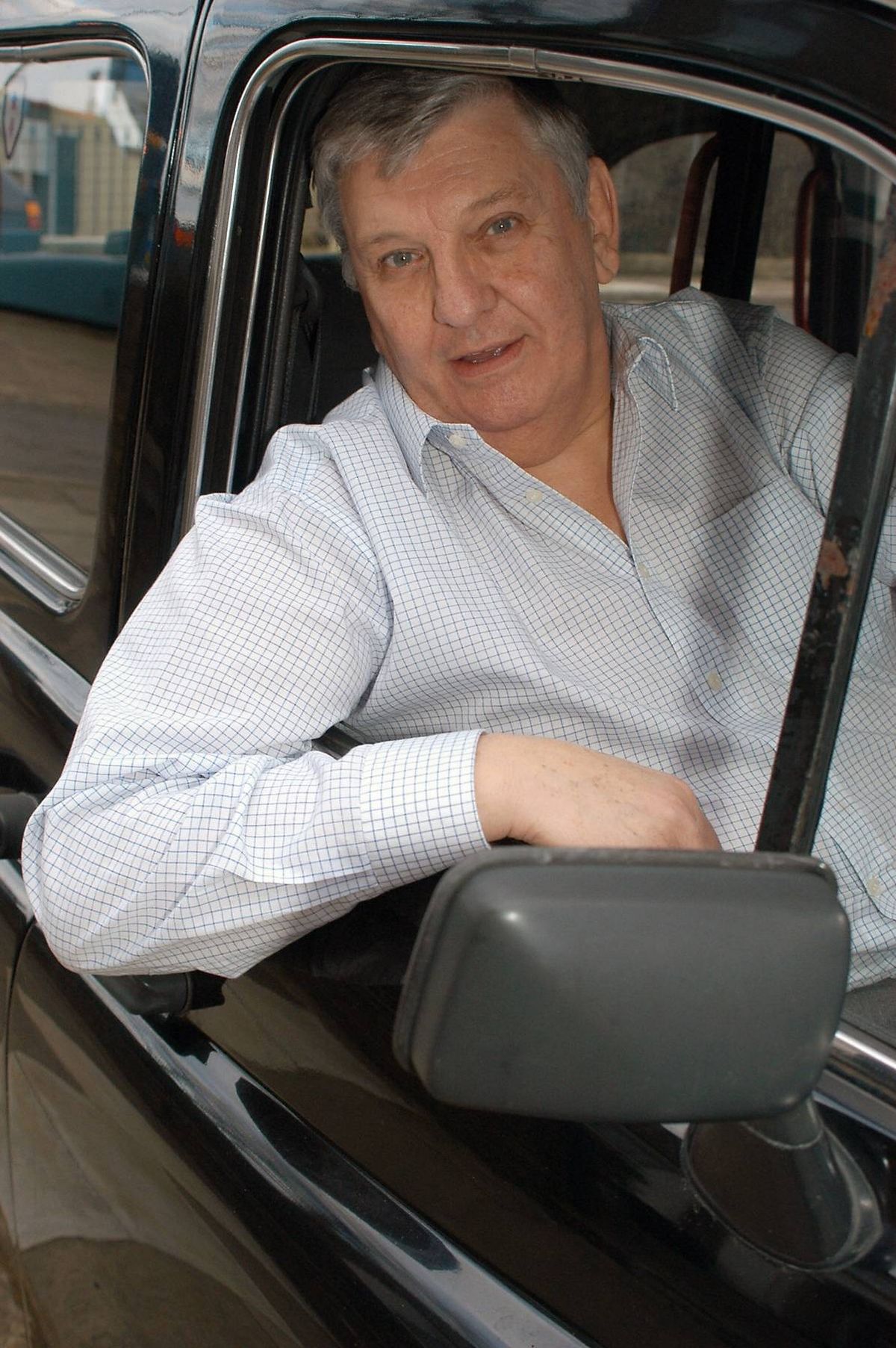 An older gentleman is seated in the passenger seat of a vehicle, wearing a light-colored shirt. The setting appears to be an outdoor environment with a blurred background, possibly indicating motion or a deliberate photographic technique.