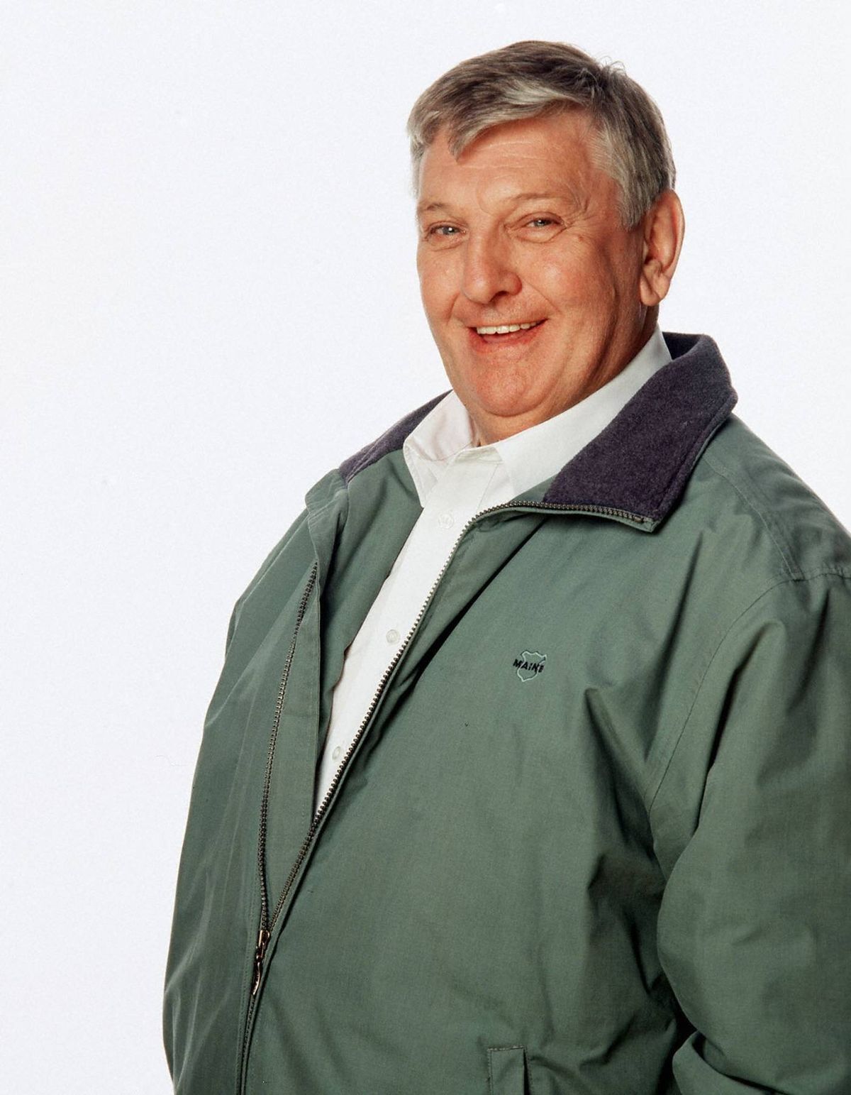 An individual with a cheerful expression is donning a white undershirt and a green jacket with a dark-colored collar. The person is positioned against a plain white backdrop.