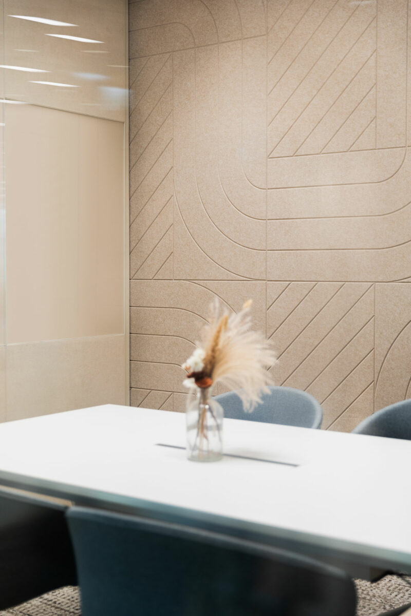 A minimalist meeting room with a white table, blue chairs, and a glass vase holding dried grasses, set against a textured beige wall with geometric patterns.