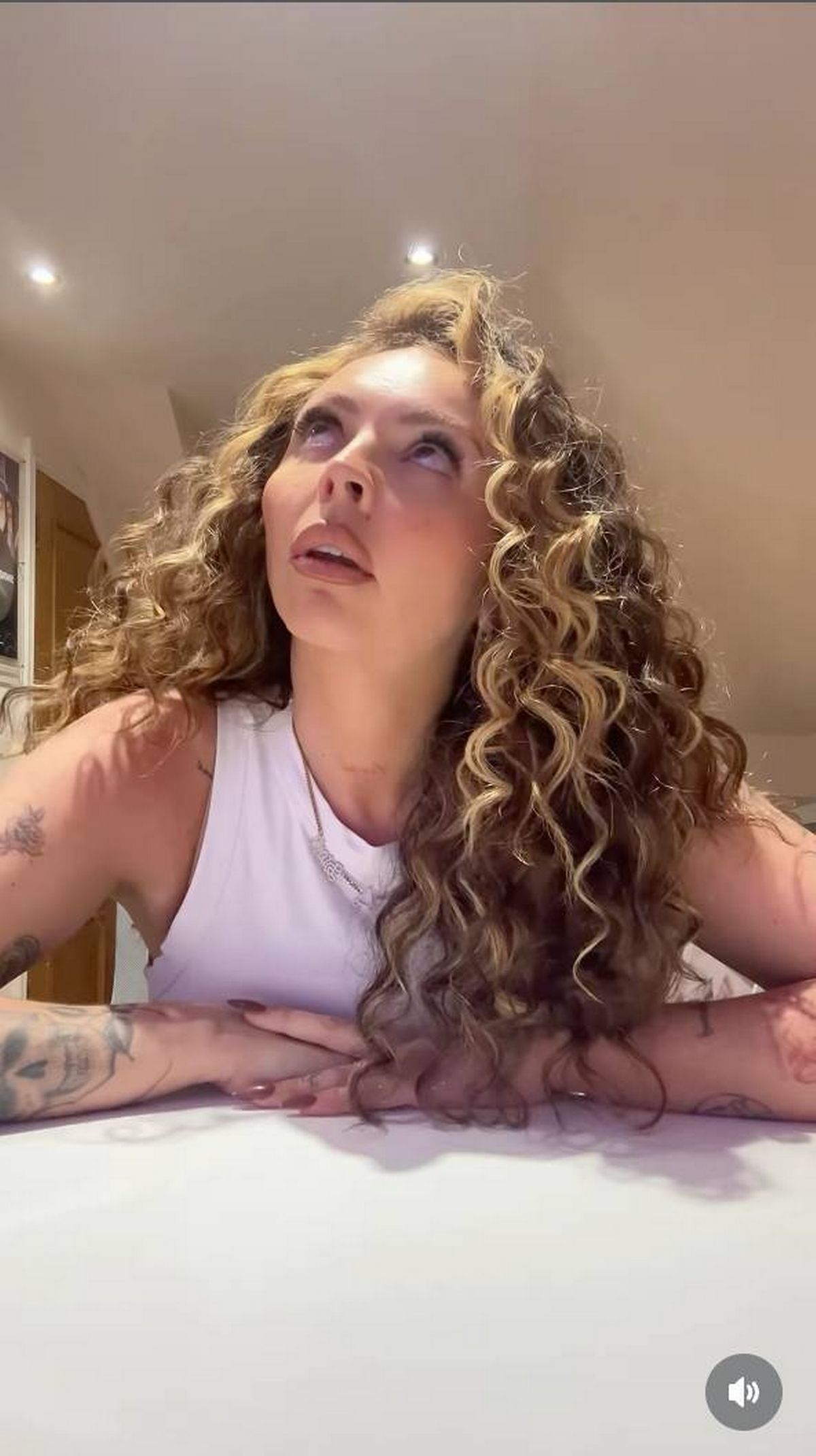 An individual with long, curly hair is seen from the shoulders up, wearing a sleeveless top. The person appears to be speaking or singing while looking upward, likely in a room with a beige wall in the background.