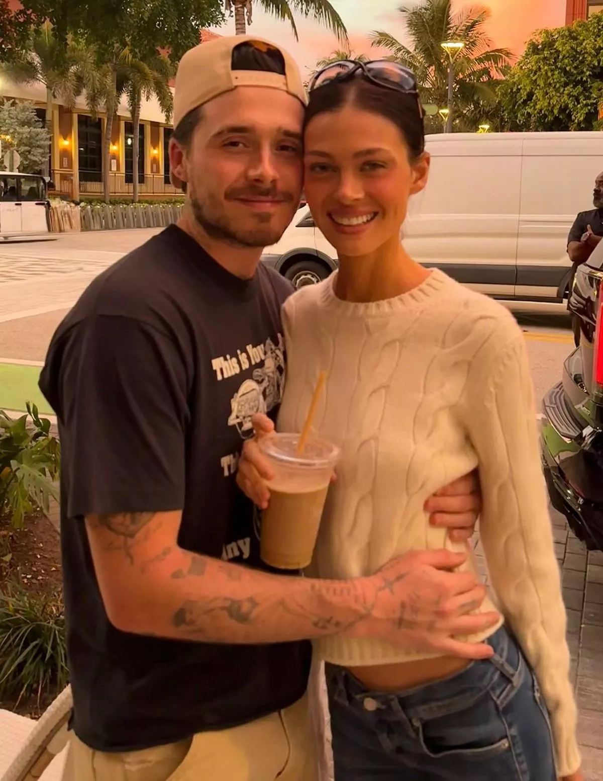 An individual in a cap and a companion are standing together, smiling, with a white van and lush greenery in the background.