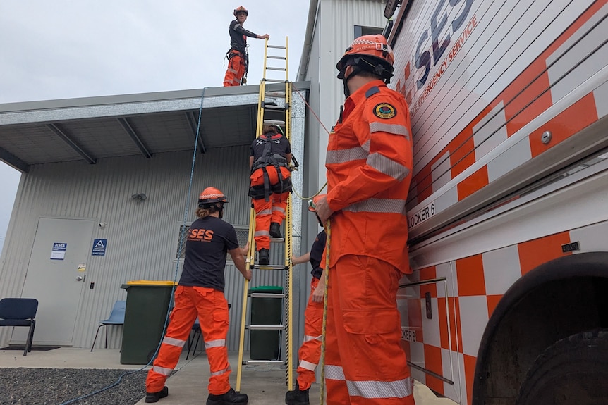 People in high-vis work with a ladder near an emergency truck and a metal-walled building.