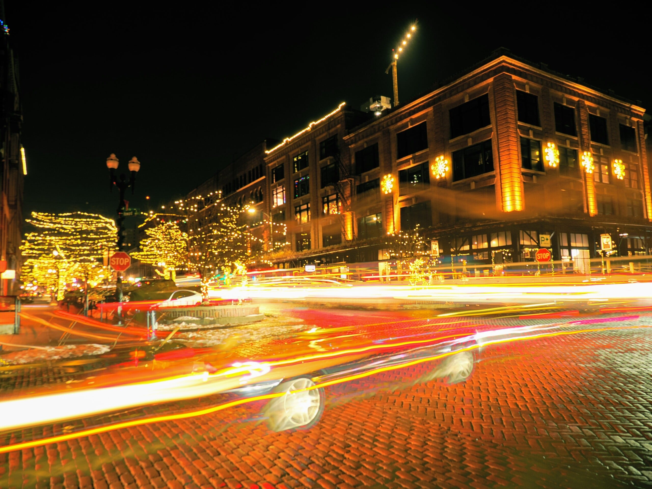 A city street at night with brick pavement, buildings decorated with holiday lights, illuminated trees, and streaks of car lights creating a sense of motion.