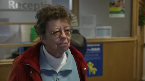A woman with glasses and a red coat stands in the reception area of the doctors surgery. She is talking to someone off camera and is staring intently. Glass windows and a desk can be seen in the background.