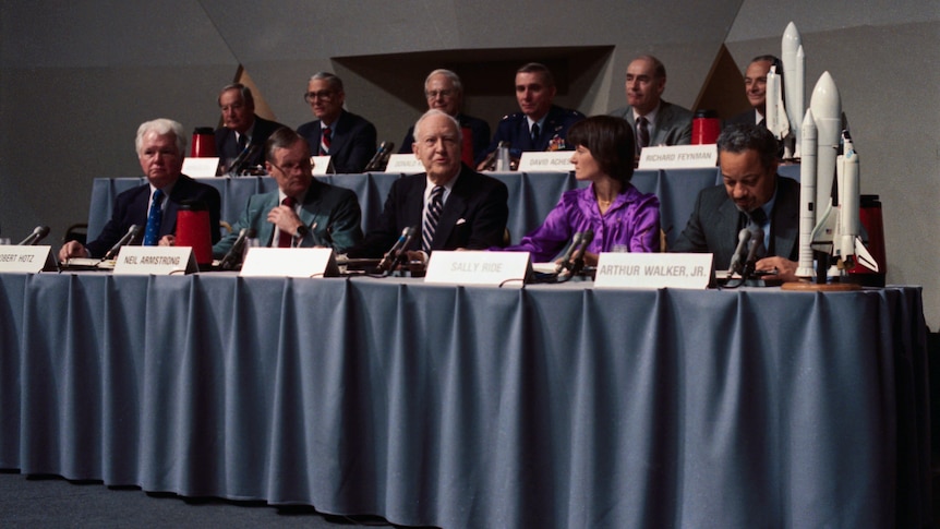 Eleven people sit formally in two rows in front of tables with nameplates, blue tablecloths and two model space shuttles.