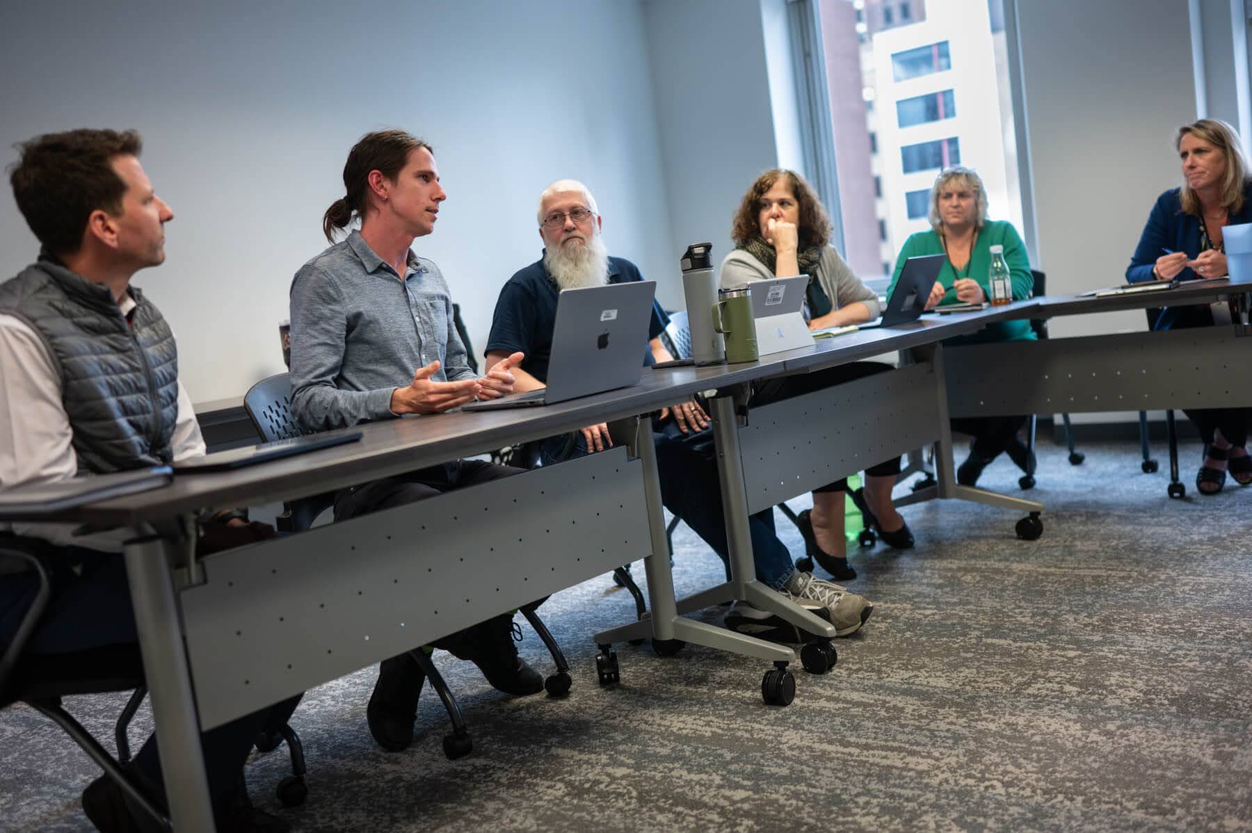 A group of six people sit around a conference table in a meeting room, engaged in discussion with laptops and notepads in front of them.
