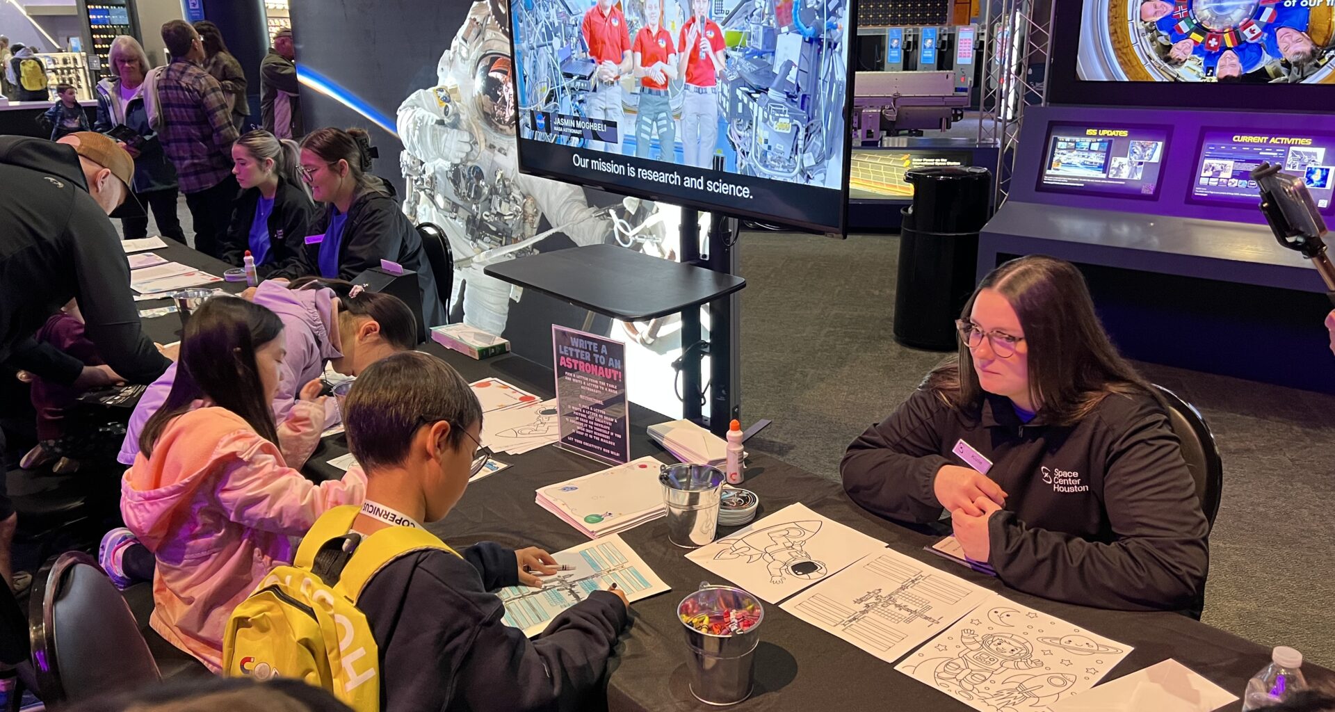 Children participate in a hands-on space activity at Space Center Houston, placing stickers on a poster at an interactive table while visitor center team members provide guidance.