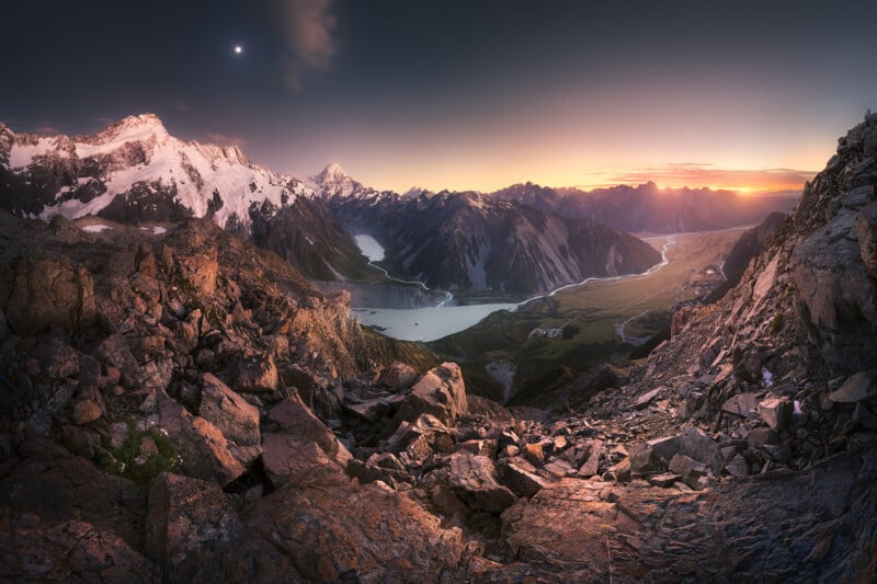 Rocky mountain landscape at sunset with snow-capped peaks, a winding river, and a lake below. Soft sunlight illuminates the rocks in the foreground; the sky transitions from dusk to night with a visible moon.