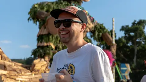 Matt Collins A man in a baseball cap and sunglasses smiles.