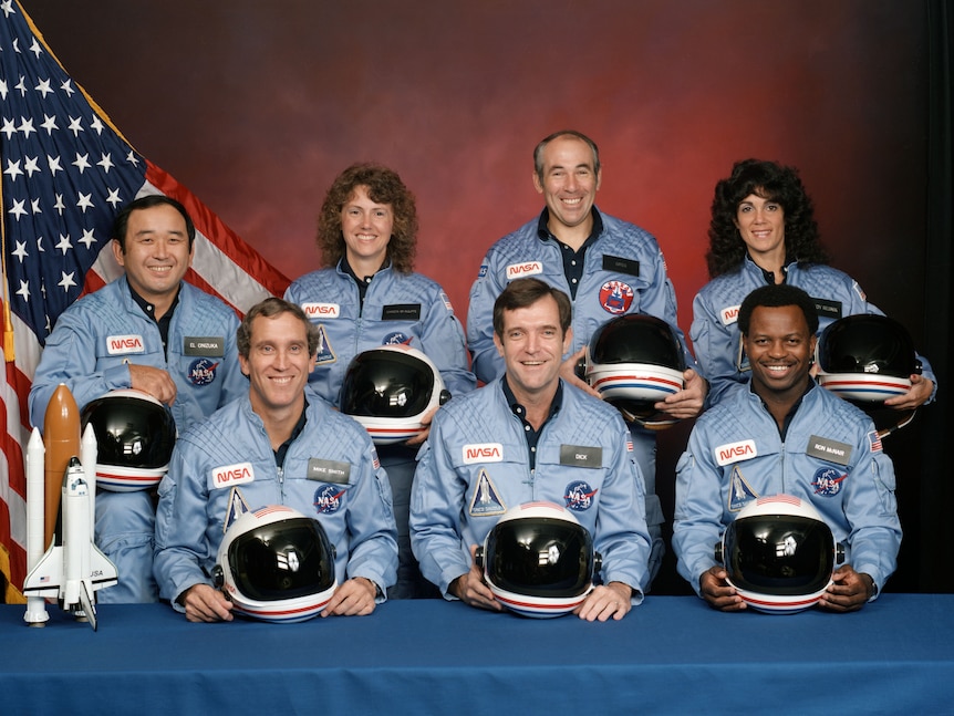 Formal studio portrait of seven astronauts wearing flight suits and holding helmets, with an American flag on one side.