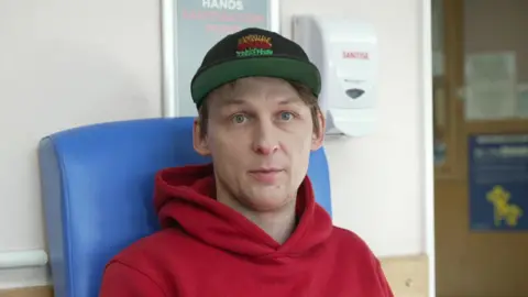 A man wearing a red hoodie and black and green baseball cap stares into the camera. He is sitting on a blue chair in a doctors waiting area. A hand sanitiser dispenser is visible in the background.
