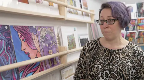 A woman with purple short hair is wearing glasses and an animal print dress. She is standing in front of her colourful print-style artwork.