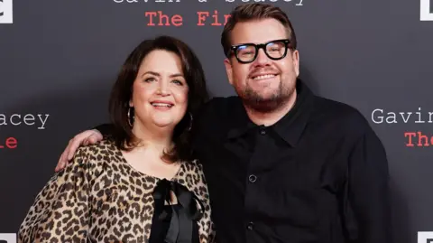 PA Media Ruth Jones is standing on the left, wearing a leopard print blouse tied with a black bow in the front. James Corden has his arm around Jones' shoulder, wearing a black shirt and thick black framed glasses