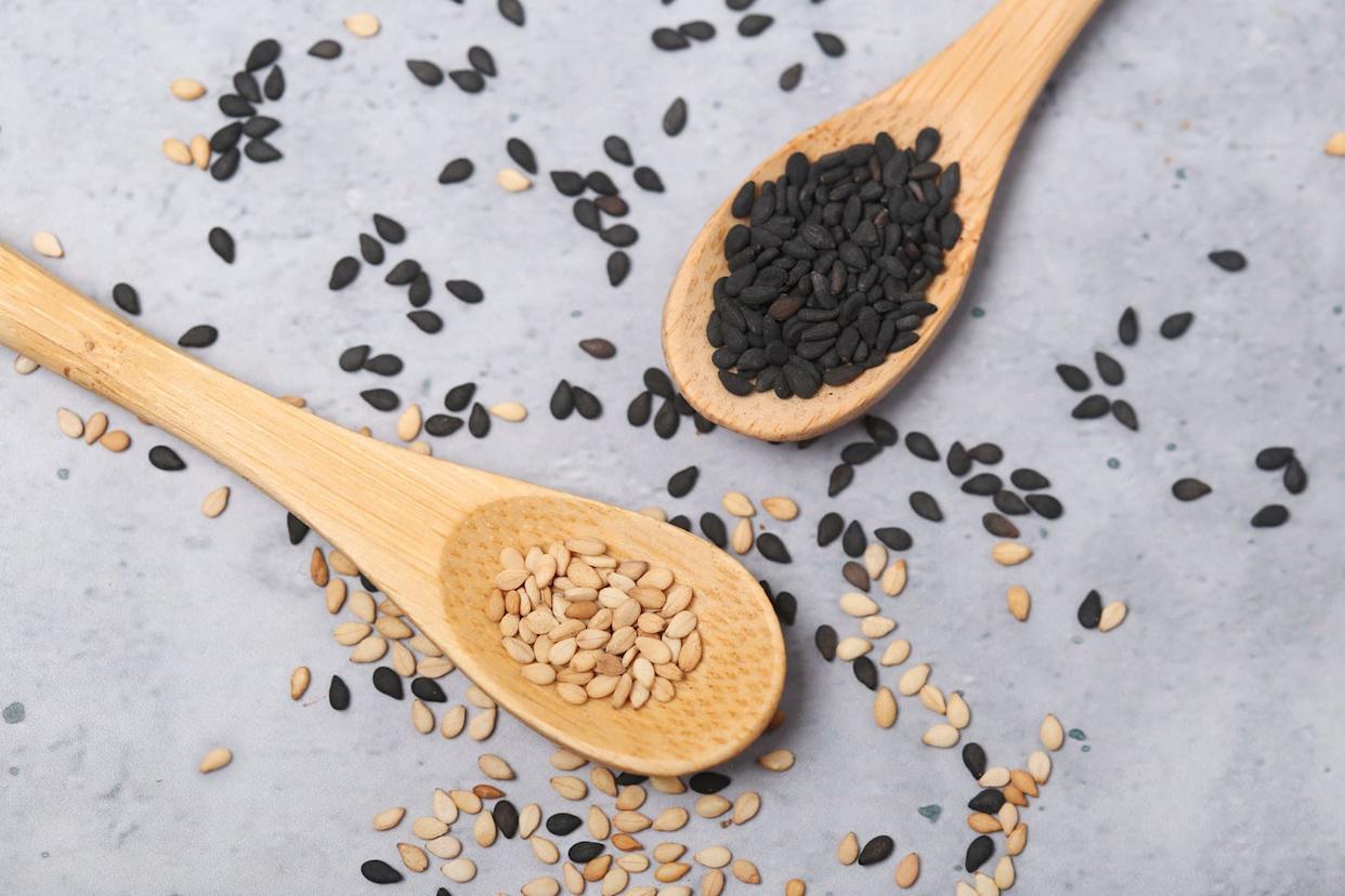 sesame seeds and black sesame seeds in wooden spoons over a gray table strewn with sprinkled sesame and black sesame seeds